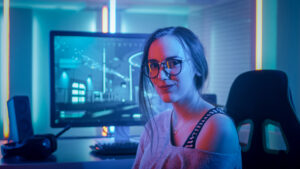 Portrait of the Beautiful Young Pro Gamer Girl Sitting at Her Personal Computer and Looks into Camera. Attractive Geek Girl Player Wearing Glasses in the Room Lit by Neon Lights.