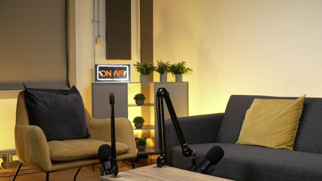 Modern studio featuring a grey couch with yellow pillows, yellow armchair, table, and two microphones against an ambient yellow backdrop.
