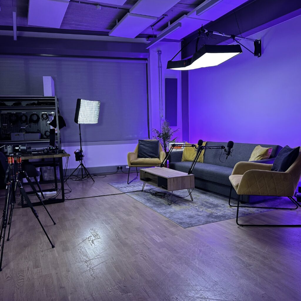 grey couch with yellow pillows, two armchairs, table, 4k cameras and two microphones against an ambient purple backdrop.