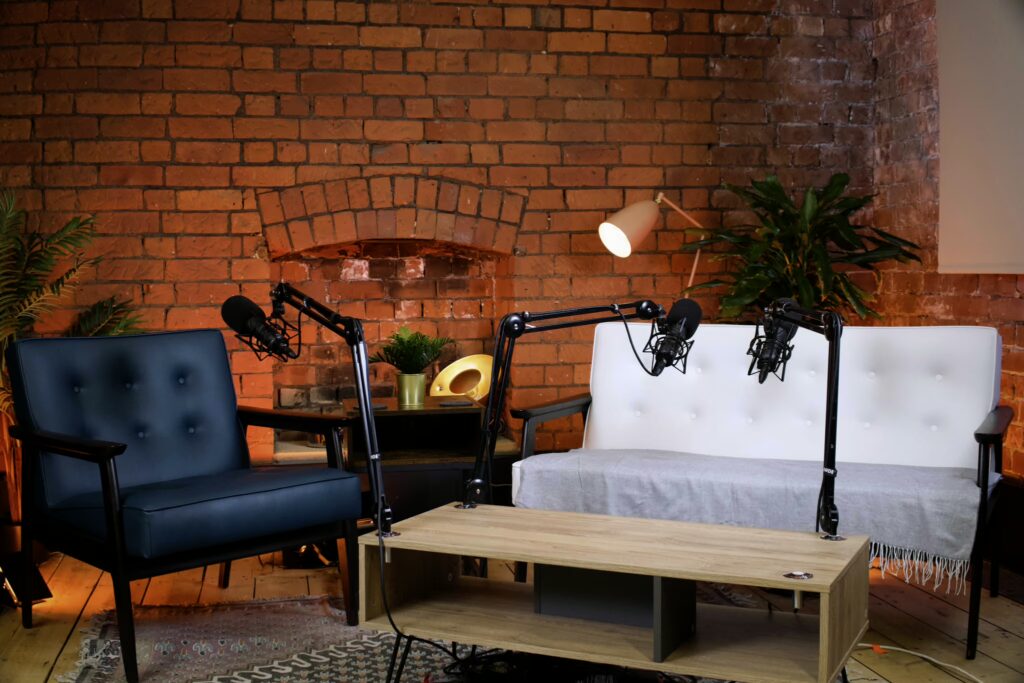 Cozy podcast studio featuring a blue accent chair, white sofa, microphones, and warm lighting against a brick wall backdrop.