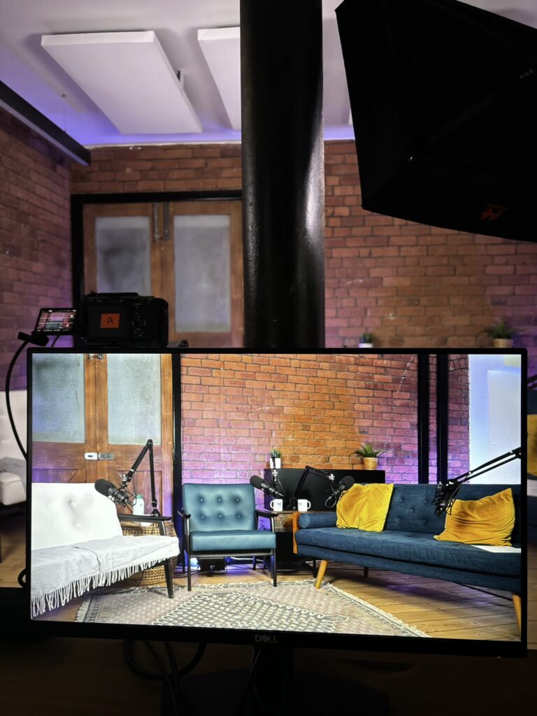 Comfortable podcast studio featuring a white sofa blue accent chair and blue sofa, and purple lighting against a brick wall backdrop.