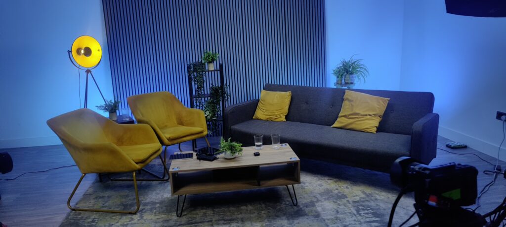 A cozy podcast studio setup featuring a gray sofa, yellow armchair, and microphones, with cameras and ambient blue lighting ready for recording.